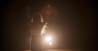 Bir kız, arabanın arka planında geceyi aydınlatan farlara karşı gölgede özgürce dans ediyor. Şehir ortamında karanlıkla çevrili neşeli hareketini yakaladığı an..