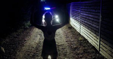Young girl stands with hands raised on a dark dirt road, facing a police silhouette illuminated by distant lights. The environment feels charged with emotion and uncertainty.