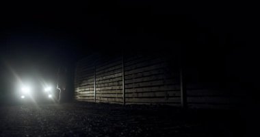 A runner moves swiftly along a shadowy trail, with distant lights casting eerie silhouettes on the surrounding environment. The atmosphere is quiet and intense.