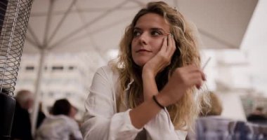 Uzun sarı saçlı genç bir kadın bir kafe masasında oturup sigara içiyor ve sohbet ediyor. Atmosfer, arka plandaki insanlarla dolu. Günlerinin tadını çıkarıyorlar..