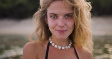 Saçları dağınık bir kadın, güneş batarken deniz kenarında huzurlu bir anın tadını çıkarır. Parlak bir şekilde gülümsüyor, deniz kabuğu kolyesi ve siyah bir mayo giyiyor, mutluluk saçıyor..