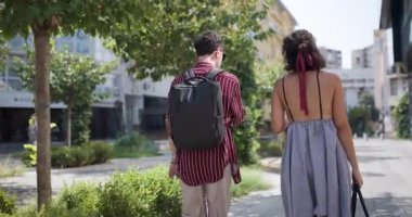Bir çift arkadaş şehir ortamında yürüyor, güneşli bir günün tadını çıkarıyor ve sohbet ediyorlar. Çevre, yeşil yapraklı, modern..