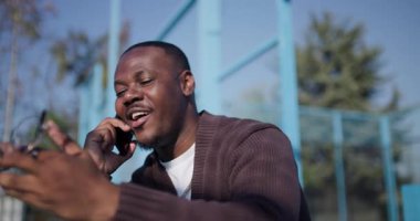 Genç bir adam açık hava spor sahasında bir bankta oturuyor, telefonuyla konuşuyor ve elleriyle işaret ediyor..