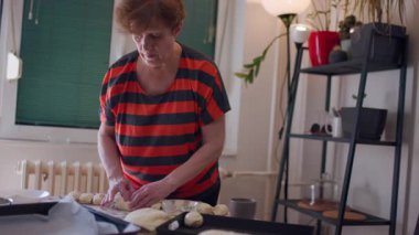 Yaşlı bir kadın mutfağında ev yapımı rulo yapıyor. O, hamuru dikkatlice şekillendiriyor ve bir pişirme çarşafına yerleştiriyor..