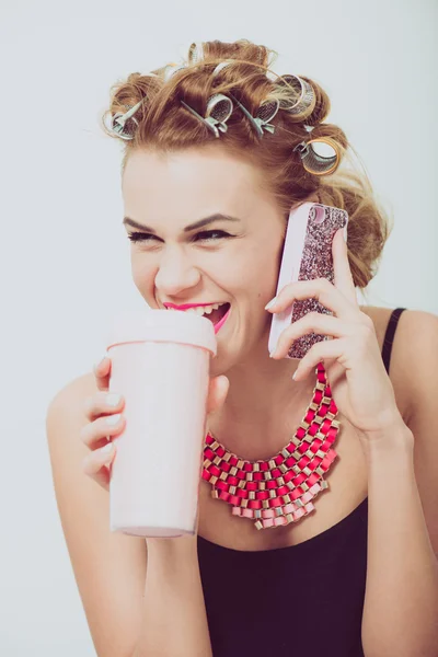 若い女性は、家で携帯電話を使用して。彼女の手と彼女の髪にカーラー飲料. — ストック写真