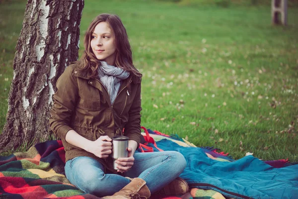 Young brunette woman resting under a tree during trip. Caucasian backpacker woman holding a mug outdoor. — Stock Photo, Image