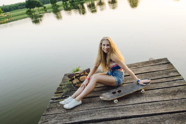 Schattig meisje rusten aan het meer met skateboard bij zonsondergang — Stockfoto