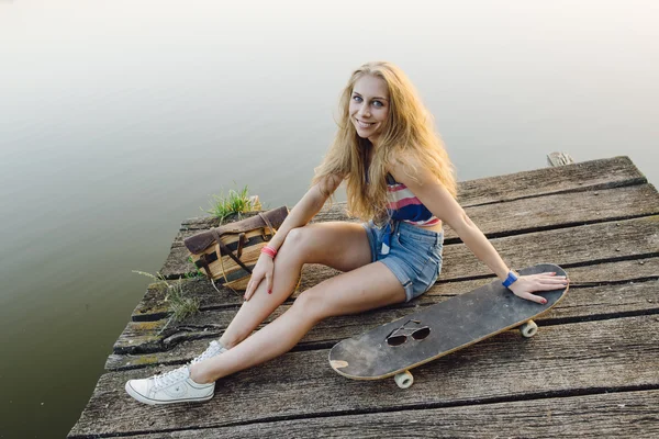 Linda chica descansando junto al lago con monopatín al atardecer — Foto de Stock