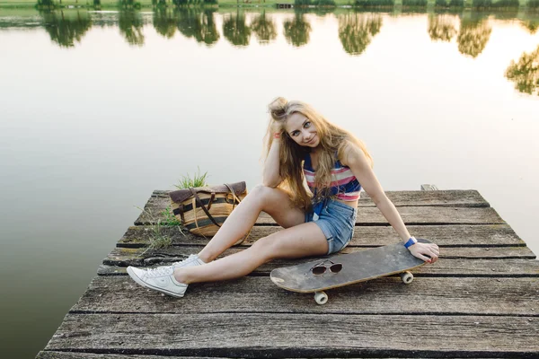 Schattig meisje rusten aan het meer met skateboard bij zonsondergang — Stockfoto