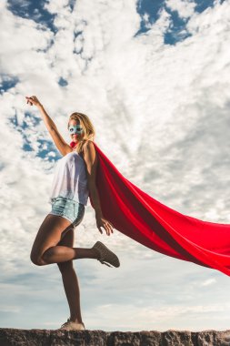 Blonde woman in red dress and red mantle posing outdoor as a superhero against blue sky clipart