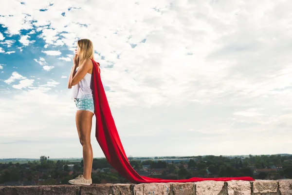 Блондинка в красном платье и красной мантии, изображающая супергероя на фоне голубого неба — стоковое фото