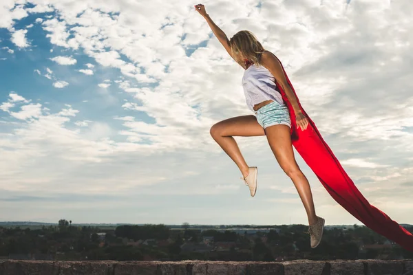 Mulher loira em vestido vermelho e manto vermelho pulando ao ar livre como um super-herói contra o céu azul — Fotografia de Stock