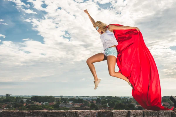Szőke nő piros ruhában és piros köpeny ugrás szabadtéri, mint egy szuperhős kék ég ellen — Stock Fotó