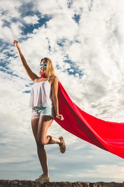 Mujer rubia en vestido rojo y manto rojo posando al aire libre como un superhéroe contra el cielo azul — Foto de Stock