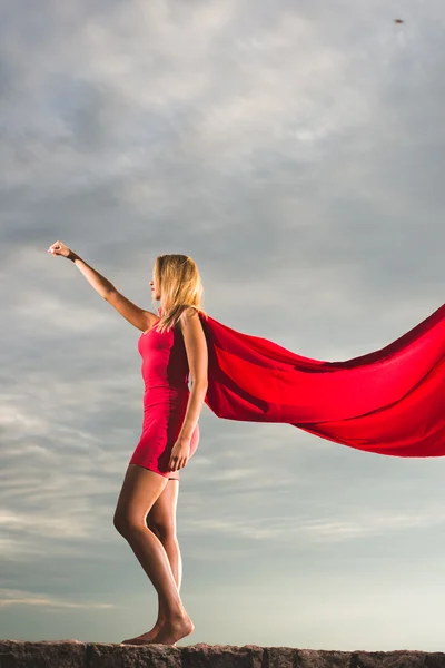 Mulher loira em vestido vermelho e manto vermelho posando ao ar livre como um super-herói contra o céu azul — Fotografia de Stock