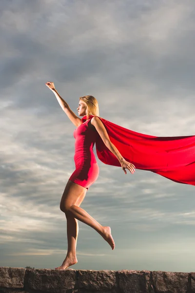 Blondýnka v červených šatech a červený plášť pózuje jako superhrdina proti modré obloze — Stock fotografie