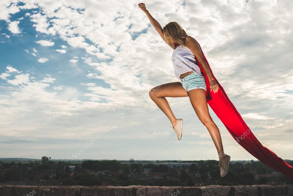 Blonde woman in red dress and red mantle jumping outdoor as a superhero against blue sky