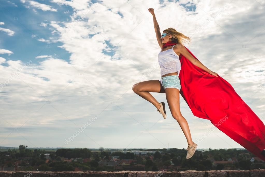 Blonde woman in red dress and red mantle jumping outdoor as a superhero against blue sky