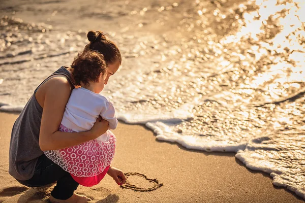 Madre e hija pequeña dibujando corazón en la playa de arena — Foto de Stock
