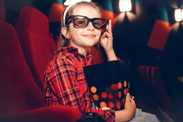 Little girl watching cartoon 3D movie in cinema and eating popcorn