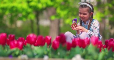 Güzel küçük kız bahar lalelerinden ve renkli çiçeklerden oluşan renkli bahçenin tadını çıkarıyor.