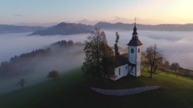 Gün batımında Slovenya 'daki Saint Thomas kilisesi ve sisle kaplı dağ tepelerinin hava manzarası