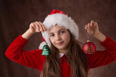 Kız el ele tutuşup Noel ağacı toplarını süslüyor ve Noel Baba şapkası takıyor, kırmızı arka planda tatil portresi çiziyor.