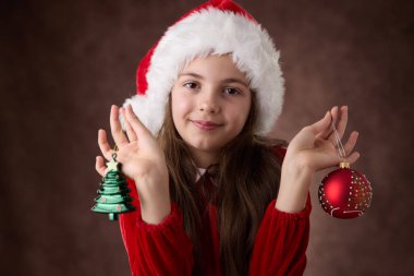 Gülümseyen kız, Noel ağacı toplarını gözlerine daya ve Noel Baba şapkası portresini kırmızı arka planda tak.