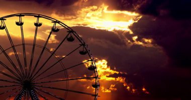 Dramatik bulutlu gün batımı gökyüzü ve lunapark karnavalında dönen dönme dolap siluetleri.