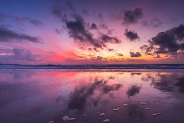 Renkli gökyüzü bulutları ve sakin okyanus dalgalarının üzerindeki yansımalarıyla sersemletici günbatımı kumsalı.