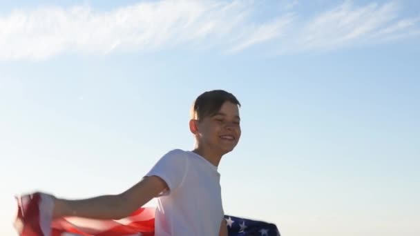 Ragazzo biondo sventola bandiera nazionale USA all'aperto sopra il cielo blu sulla riva del fiume — Video Stock