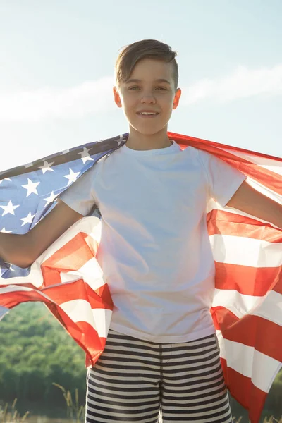 Blonďatý chlapec mává národní vlajkou USA venku nad modrou oblohou na břehu řeky — Stock fotografie