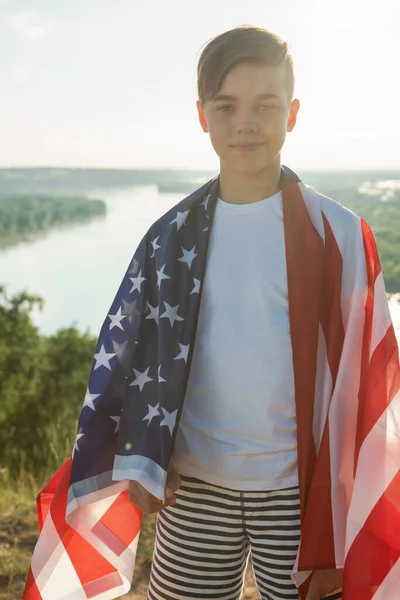 Niño rubio ondeando bandera nacional de EE.UU. al aire libre sobre el cielo azul en la orilla del río —  Fotos de Stock