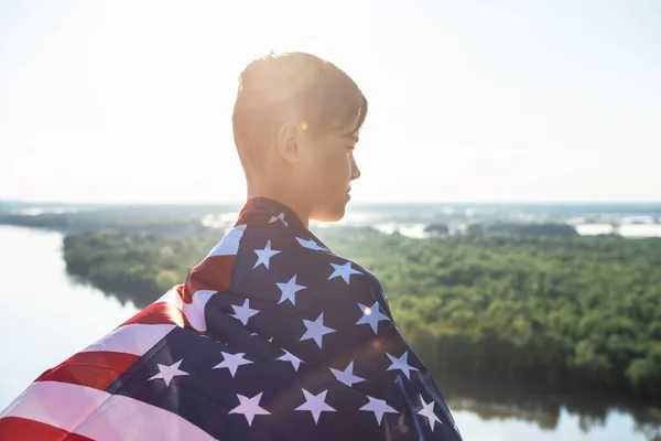 Blond pojke viftar nationell USA flagga utomhus över blå himmel vid floden banken — Stockfoto