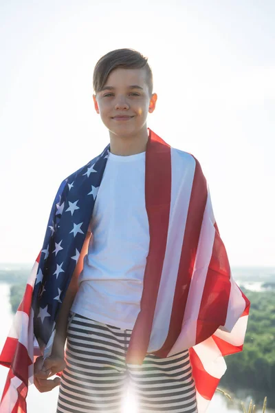 Blonďatý chlapec mává národní vlajkou USA venku nad modrou oblohou na břehu řeky — Stock fotografie