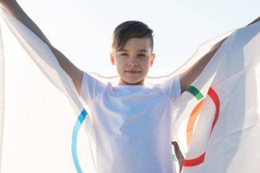 Sarışın çocuk nehir kıyısında açık havada Olimpiyat Oyunları bayrağını sallıyor.