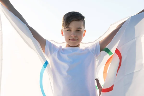 Niño rubio ondeando bandera los Juegos Olímpicos al aire libre sobre el cielo azul en la orilla del río —  Fotos de Stock