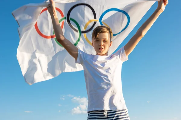Blonde boy waving waving flag the Olympic Games outdoors over blue sky at the river bank — Stock Photo, Image