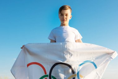 Sarışın çocuk nehir kıyısında açık havada Olimpiyat Oyunları bayrağını sallıyor.