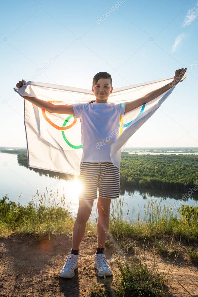 Portrait of boy waving flag the Olympic Games outdoors over cloudy sunset sky. Children sports fan. Summer olympic games concept. 08.06.2021, Barnaul Russia.