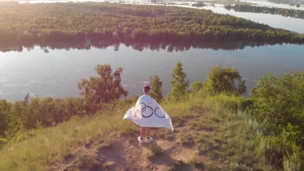 Luftdrohne 4k Video von Junge, der Flagge der Olympischen Spiele über blauem Himmel am Flussufer schwenkt — Stockvideo
