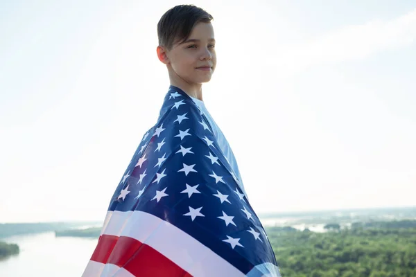 Blonde boy waving national USA flag outdoors over blue sky at the river bank — ストック写真