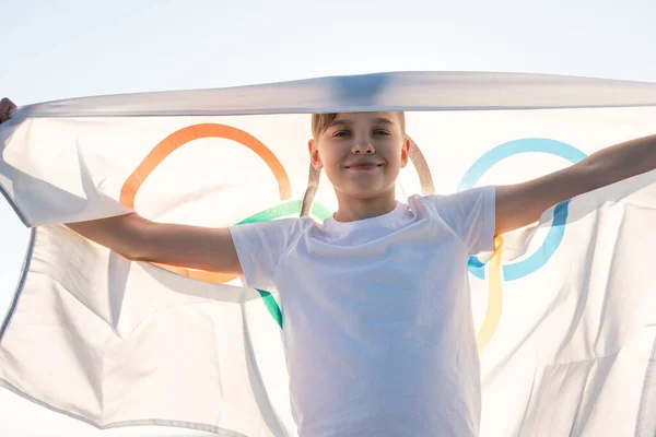 Blonde boy waving waving flag the Olympic Games outdoors over blue sky at the river bank — Stock Photo, Image