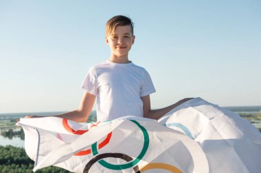 Sarışın çocuk nehir kıyısında açık havada Olimpiyat Oyunları bayrağını sallıyor.