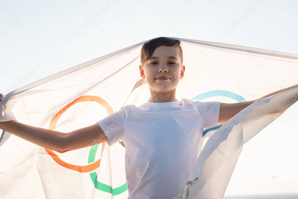 BARNAUL CITY. RUSSIA - JUNE 08, 2021: Portrait of boy waving flag the Olympic Games outdoors over cloudy sunset sky. Children sports fan. Summer olympic games concept on June 08, 2021 in Altayskiy