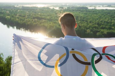 Sarışın çocuk nehir kıyısında açık havada Olimpiyat Oyunları bayrağını sallıyor.