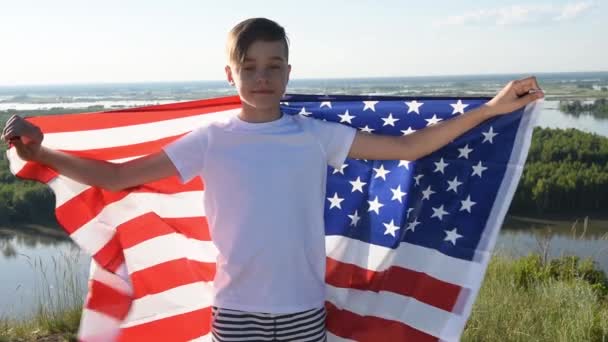 Blonďatý chlapec mává národní vlajkou USA venku nad modrou oblohou na břehu řeky — Stock video