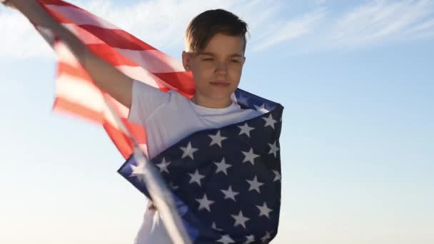 Niño rubio ondeando bandera nacional de EE.UU. al aire libre sobre el cielo azul en la orilla del río — Vídeos de Stock