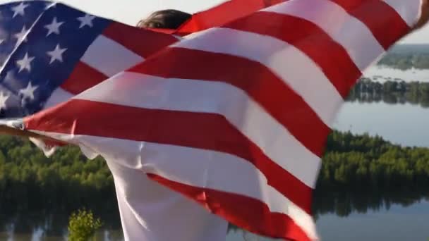 Блондин, размахивающий национальным флагом США над голубым небом на берегу реки — стоковое видео