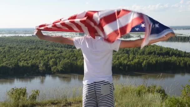 Blonďatý chlapec mává národní vlajkou USA venku nad modrou oblohou na břehu řeky — Stock video